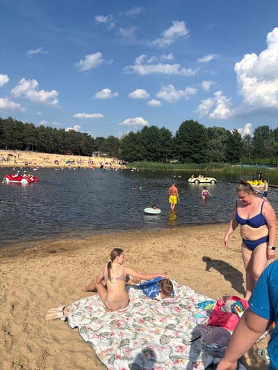 The image depicts a sunny day at a lake beach in Kobyla Góra, Poland. A sandy shore is in the foreground with several people relaxing on a blanket. In the water, there are multiple individuals swimming, sunbathing on inflatables, and using paddle boats. A red paddle boat shaped like a car is visible on the left, and a yellow pedal boat resembling a car is in the middle of the lake. The background shows a dense forest bordering the lake. The sky is blue with scattered white clouds.