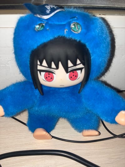 A stuffed toy resembling the anime character Sasuke Uchiha from Naruto, wearing a blue, fuzzy animal costume, sits on a wooden surface. The toy has black hair, a pale face, and red eyes with the Sharingan pattern. Its body is covered in blue fur, with small, tan feet visible at the bottom. Two black cables are present in the foreground, partially obscuring the toy's feet. The background is a white, brick-patterned surface. There is no information to suggest the location is in Melitopol, Ukraine.