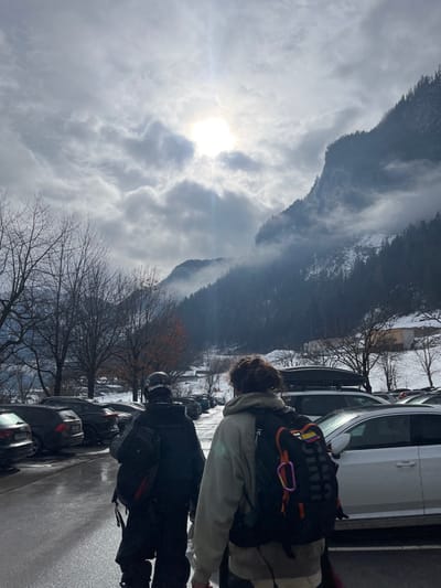 A vertical shot shows people and cars alongside a road, with a mountainous background. People with backpacks walk on a wet pavement. Cars are parked to the right of the road. Beyond the road, bare trees line the edge. The sky is partly cloudy, with the sun shining through. A large mountain looms in the background, partly covered in snow.