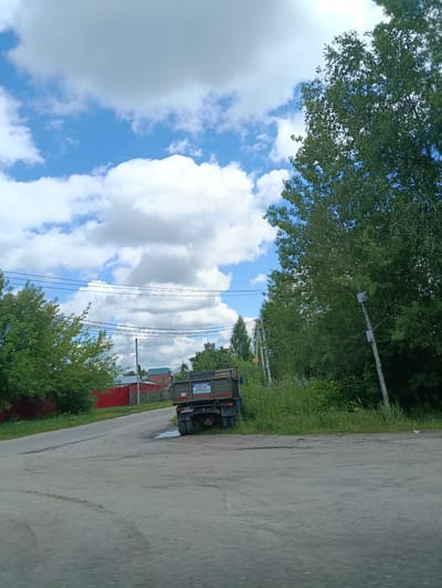 The image depicts a rural road scene in the settlement of Тополиный, Russia, on a bright, cloudy day. The road, made of asphalt, curves to the left and appears somewhat weathered, with some tire marks visible. On the right side of the road, tall green trees and dense vegetation line the area. A single utility pole stands near the roadside. In the distance, to the left of the road, a red fence encloses properties with red and green roofed buildings.
Parked on the right side of the road, partially on the shoulder, is a green flatbed truck. The truck has dual rear wheels and a license plate that is partially obscured but appears to read "PA5869SS." A blue and white sticker or sign is visible on the side of the truck's bed. A puddle of water is present on the road near the truck. Overhead, power lines are strung across the scene, supported by poles. The sky is a mix of bright blue and large, puffy white and grey clouds.