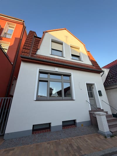 The image displays a street view in Rostock, Germany, featuring the facade of a multi-story residential building. The building has a light-colored stucco exterior with a red-tiled, steeply pitched roof. There are several windows visible, including a large, multi-pane window on the ground floor reflecting a sailboat model. Above this, on the upper level, are two smaller windows with roll-down shutters. A gray-painted door is located to the right of the ground-floor window, accessible by a set of granite steps. A green mailbox is mounted to the right of the door. The foreground shows a paved sidewalk made of reddish-brown bricks and gray cobblestones. Adjacent buildings, one painted bright orange, are also partially visible, creating a dense urban setting. The sky is clear and blue, with the sun casting warm light on the building.