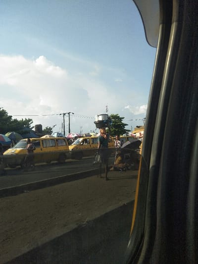 The image, captured from inside a vehicle looking out the passenger-side window, depicts a busy street scene in Awjaw, Nigeria. The foreground shows a dark, unpaved ground surface leading to a raised curb, beyond which lies a paved road. The sky is largely clear blue with scattered white clouds.
In the mid-ground, a female individual walks towards the viewer's left on the paved road, balancing a stack of various containers, including metallic pots and plastic bowls, on her head. She is dressed in a striped top and a dark skirt, carrying a bag in her left hand. To her right, another individual is seated on the ground beneath a dark, open umbrella.
Multiple yellow passenger vans are visible on the paved road, appearing to be stopped or moving slowly. Several colorful striped market umbrellas are interspersed among these vehicles, suggesting active street vendor stalls. Additional people are present near the vans and market areas. Utility poles with overhead wires are visible across the mid-ground, with green trees and indistinct structures in the background. The vehicle's dark A-pillar and door frame occupy the right side of the frame.