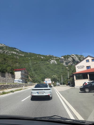 The image shows a view from a vehicle driving on a road next to a steep, forested mountainside. A white Porsche SUV is in front, driving on the same road. To the left of the road is a building with the name "ISTOK RESTORAN" visible. Further up the mountainside, more buildings and power lines can be seen. The sky is clear and blue, suggesting a sunny day. The scene appears to be in Lapčići, Montenegro. The time of day is likely midday due to the bright sunlight and clear sky. The overall impression is that of a scenic drive through a mountainous region.