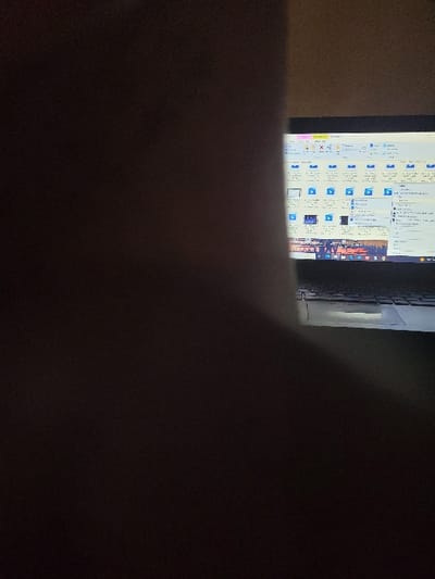 The scene, stated to be located in Kubare, Nigeria, depicts a dimly lit interior environment. The right portion of the image displays a partially visible laptop computer, with its screen illuminated. The screen shows a graphical user interface, likely a file explorer, featuring rows of light blue icons and small thumbnail previews arranged in a grid. Below these, a horizontal section of the screen displays what appears to be a still frame or image, possibly depicting a night scene with multiple artificial light sources, resembling a cityscape or a crowded event. A segment of the laptop's keyboard is visible below the screen. The left and central portions of the image are dominated by a large, amorphous dark brown to black mass, obscuring a significant portion of the foreground and left side of the laptop. No individuals are clearly identifiable, and no direct actions or interactions beyond the display on the laptop screen are discernible.