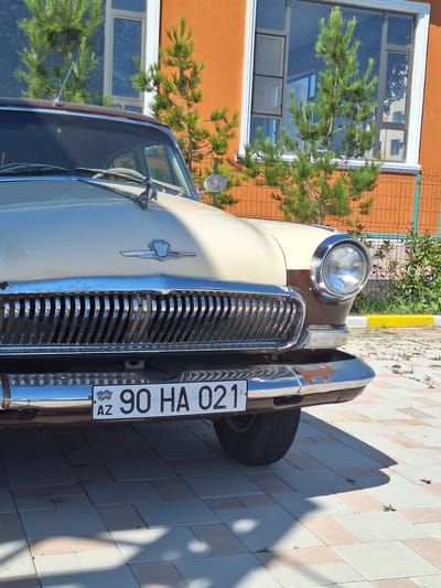 This is a close-up, eye-level, outdoor, medium shot of the front of a vintage cream and brown car. The car's prominent chrome grille dominates the lower half of the image, with vertical slats reflecting light. A license plate reading "AZ 90 HA 021" is mounted on the lower bumper. The car has a large, round headlight with a chrome bezel on the right side. The background features an orange building with a large window, framed by white trim. Several small evergreen trees are planted in front of the building, with a metal fence visible behind them. The ground is paved with light-colored interlocking bricks, and shadows from the car and trees are cast across the surface. The scene appears to be a sunny day, possibly in the city of Khudat, Azerbaijan, given the license plate format.