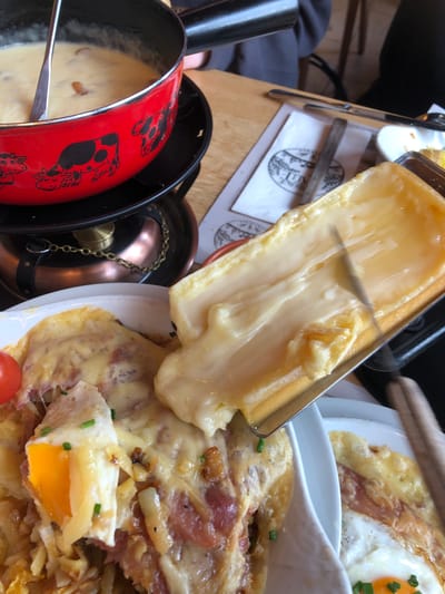 The image shows a close-up view of a dining table. Raclette cheese is being scraped from a metal holder onto a plate of food. The dish on the plate includes potatoes, bacon, and a fried egg, topped with melted cheese. In the foreground, there's a red fondue pot with a cow design. The dining table appears to be located in a restaurant or similar setting. The specific location is Epagny, Switzerland.