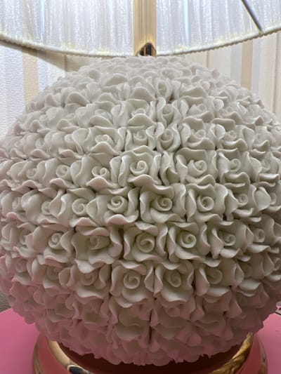 The image shows a close-up, low-angle shot of a spherical white ceramic lamp base decorated with numerous small, intricately detailed rose-shaped carvings. The lamp base is positioned on a pink surface. In the background, a portion of a lampshade with a ribbed texture and a faint vertical striped pattern on the wall are visible. The overall scene appears to be indoors, in a domestic setting. No specific location in Talgar, Kazakhstan can be determined from the visual information provided.