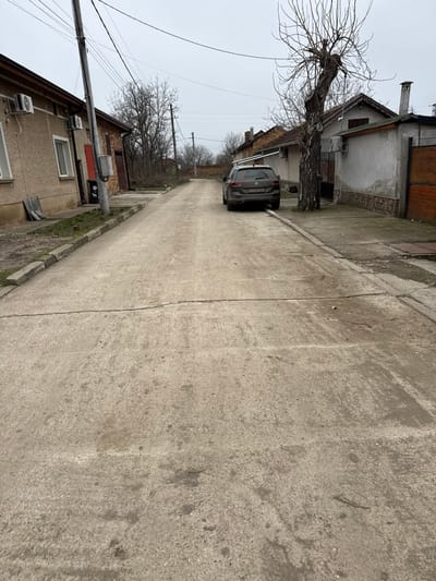 The scene depicts a narrow, concrete-paved street in Gorna Mitropolia, Bulgaria, under an overcast sky. The street surface is discolored and exhibits multiple cracks. Looking away from the viewer, the street appears to ascend gradually.
On the left side of the street, a light-brown, two-story building is visible, featuring two windows and two exterior air conditioning units. A red door is partially visible to the right of the windows. A dark gray utility pole with multiple overhead power lines stands in front of this building, adjacent to a raised, curbed dirt verge.
On the right side of the street, a dark gray Volkswagen Passat Variant station wagon (license plate CA 6100 HC) is parked parallel to the curb, facing away from the viewer. A tow hitch is visible at the rear of the vehicle. Immediately behind the parked car, a large, gnarled deciduous tree with bare branches and exposed roots stands beside a concrete sidewalk. Further along the right, a single-story building with white stucco walls, an exterior air conditioning unit, and a visible chimney is present, followed by a section of a wooden fence.
In the background, beyond the immediate structures, additional bare trees, residential buildings, and a brick wall are visible. No individuals are present in the scene.