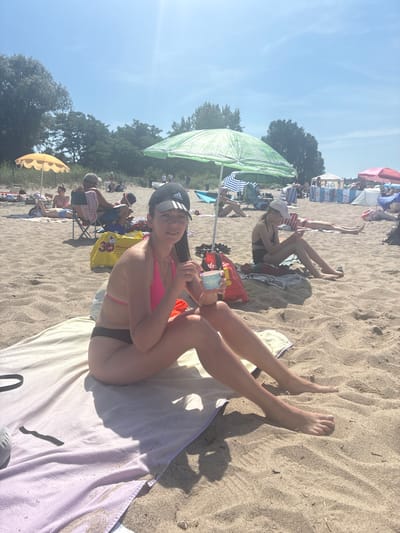 The image depicts a crowded beach scene on a sunny day in Rewa, Poland. The foreground features a young woman in a pink bikini and a black baseball cap, sitting on a light purple towel. She is holding a small blue cup and looking at the camera with a slight smile. Behind her, other beachgoers are visible, some reclining under umbrellas, others sitting in chairs. Beach umbrellas, some green and some yellow, are scattered across the sand. The sand is light brown and appears somewhat dry. In the background, lush green trees line the edge of the beach, and the sky is a clear, bright blue, suggesting pleasant weather. The overall atmosphere appears relaxed and leisurely, indicative of a typical summer day at the beach. A yellow shopping bag with red lettering is visible near the woman.