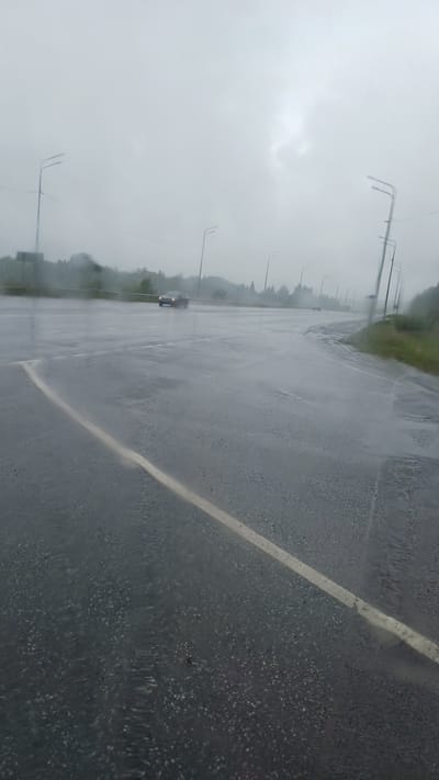 The image depicts a wet, overcast day on a road in Buranovo, Russia. The road is slick with rain, and puddles are visible on the asphalt. A white line marks the edge of a lane, and a few cars are driving on the road, their headlights illuminated. In the background, trees and streetlights line the sides of the road. The sky is a uniform gray, indicating heavy cloud cover.
