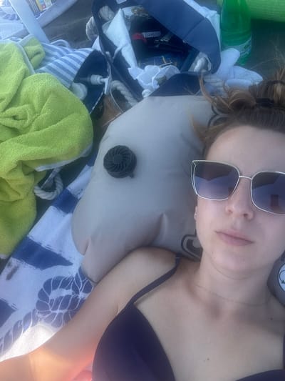 The image shows a close-up, overhead shot of a woman lying on a beach towel at Atiya, Bulgaria. She is wearing a dark purple bikini top and large, silver-rimmed sunglasses. Her hair is pulled up in a messy bun, and she is looking towards the camera. In the background, there are several beach items, including a bright green towel, a blue and white striped towel, and a blue beach bag. An inflatable pillow is positioned under her head. The overall scene suggests a relaxing day at the beach.