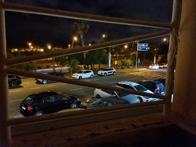 This is a nighttime, medium-distance photograph taken from a slightly elevated perspective, looking out over a street and parking area through a balcony railing. The scene is illuminated by streetlights and the headlights of vehicles.

In the foreground, a blurred brown railing runs horizontally across the frame, with a vertical tan post on the right side.

Beyond the railing, a paved street is visible with multiple cars parked and driving. To the left, a black hatchback is parked closest to the foreground. Further down the street, a blue sedan is in motion.

In the middle ground, several cars are parked in a lot or along the street. White vehicles are prominent, including two white compact cars and a white SUV. A dark-colored SUV is also visible.

In the background, there is a road with traffic signs. One sign indicates directions to "Costa Adeje," "La Caleta," and "Fañabé," and includes the road number "70" and "500 m." Palm trees and other foliage line the street. Buildings with illuminated windows are visible in the distance, suggesting an urban or suburban environment. The sky is dark, indicating nighttime. No people are clearly visible in the image.