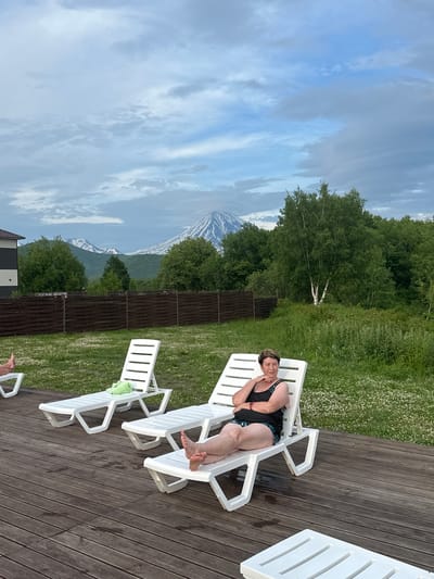A woman is lying on a white plastic chaise lounge on a wooden deck. She is wearing a black swimsuit with a patterned trim and has her head resting on her hand, with her eyes closed. The deck is surrounded by lush green grass with scattered white wildflowers. In the background, a wooden fence separates the grassy area from a dense forest of trees. Beyond the trees, snow-capped mountains rise under a cloudy sky with patches of blue. The scene appears to be set during the daytime, with the weather looking overcast. The overall impression is one of relaxation and tranquility in a natural setting.