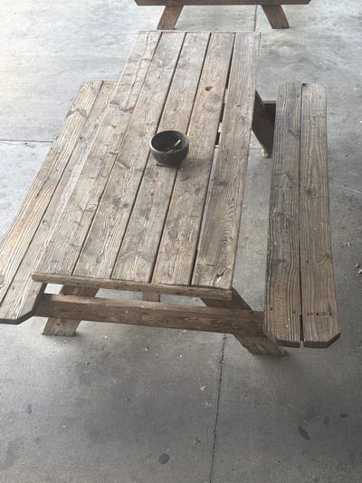 An overhead shot shows a weathered, wooden picnic table with attached benches on a gray concrete surface. The table is positioned diagonally in the frame, with the left side of the image showing more of the table and the right side showing a bench. The wood of the table and benches displays prominent grain patterns and has a distressed, aged appearance. A small, dark ashtray sits on the tabletop near the center, containing a few cigarette butts. The concrete ground around the table is uneven and shows some minor stains and debris. The lighting suggests it is daytime, possibly outdoors. The context for this scene within Murfreesboro, United States of America is not determinable from the image alone.