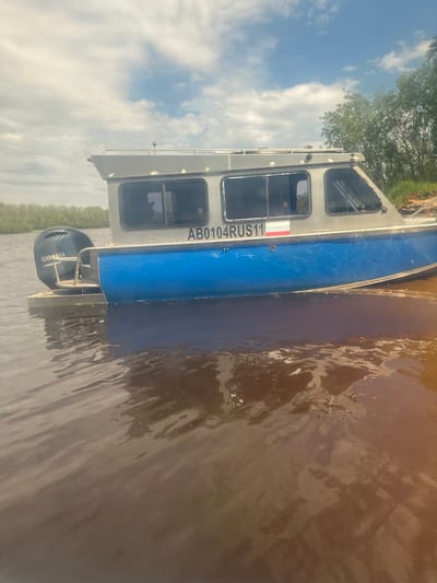 A motorboat with a blue hull and a grey cabin is docked on murky, brown water. The boat has the registration number "AB0104RUS11" and a Russian flag decal on its side. A Yamaha outboard motor is attached to the stern. The boat is situated near a vegetated shoreline, with trees visible in the background under a cloudy sky. The scene appears to be a waterfront area in the city of Ozyorny, Russia.