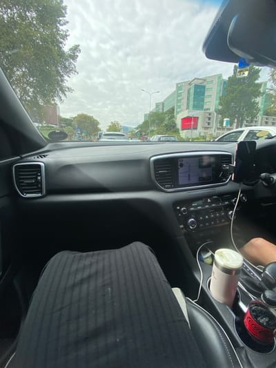 The scene is observed from the front passenger seat of a vehicle positioned on a multi-lane urban road in Sandton, South Africa. The vehicle's dark dashboard with chrome-trimmed air vents is prominent. A central infotainment screen displays navigation, and a smartphone is secured in a mount to its right. Controls for climate and media are visible below. The driver's right arm is partially visible on the far right, near the gear shift. In the center console, a light-colored thermal mug and a red Coca-Cola branded container are situated in cup holders, accompanied by white charging cables. The passenger's legs, covered by dark, vertically striped fabric, occupy the lower left foreground.
Through the windshield, an overcast sky is visible above the road. Multiple vehicles are present in traffic ahead, indicating a typical urban flow. On the right, the multi-story AON Building, characterized by its modern design and green-tinted glass, features the 'AON' logo on its facade and a large red rectangular sign below. Trees line both sides of the street, and traffic lights are visible in the distance. The vehicle is located on Sandton Drive, facing eastward, with the AON Building at 1 Sandton Drive visible on the right.