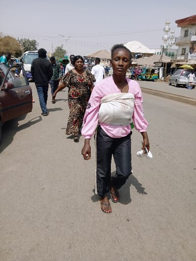 The image depicts a daytime street scene populated by multiple individuals and vehicles under a hazy sky.
In the foreground, a woman with dark skin and dark, pulled-back hair walks directly towards the viewer. She wears a pink long-sleeved top, dark trousers, and dark flip-flops, displaying red painted toenails on her right foot. A light-colored, possibly white, fabric is wrapped across her chest and torso. Her left hand is slightly clenched, holding a crumpled white object.
Behind her, slightly to the left, a second woman with dark skin walks in the same direction, wearing a long, patterned dress featuring shades of brown, black, and tan. Further back and to the left, a man wearing a dark long-sleeved shirt and blue jeans walks away from the viewer. To his immediate left, a dark-colored vehicle with its passenger door open is partially visible.
The mid-ground features several other indistinct individuals walking on the paved road. Vehicles in the background include a white minibus or van on the left, several multi-colored auto-rickshaws (tuk-tuks) on the right, and a silver sedan parked further down the street on the right.
The background consists of various buildings, some with flat roofs and others with multiple stories. On the far right, a building with distinctive white minaret-like structures is visible, suggesting a mosque or similar architectural style. The sky is light and hazy, and sparse trees are visible on the left side of the street. Overhead power lines and streetlights are present.
The visual evidence within the image does not provide information to confirm the specific location as Kungulu School, Zambia. The scene is consistent with a busy urban or semi-urban street environment in an African city.