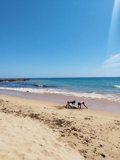This image captures a bright, sunny day at a beach in Rota, Spain, likely Playa de la Costilla given the characteristic long, sandy expanse and the presence of a breakwater. Three children are engaged in activities on the sand near the water's edge. One child, wearing a colorful swimsuit, is lying on their stomach, facing away from the camera, appearing to interact with the sand. Two other children are positioned behind them, also bent over and seemingly playing or digging in the sand. The ocean stretches out beyond them, with gentle waves lapping at the shore and a clear blue sky overhead. In the distance, a breakwater made of large stones protrudes into the sea, a common feature on Rota's coastline. The sand is light brown and shows some scattered patches of seaweed. The overall scene is one of a relaxed, family-friendly beach day.