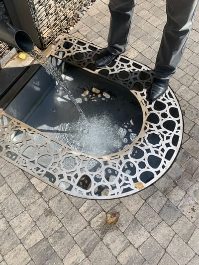In Arbuzov-Baran, Russia, a metal pipe directs a stream of water into an ornamental basin on a paved stone walkway. The basin is surrounded by a decorative metal grate with an intricate geometric pattern. Two individuals, dressed in dark pants and shoes, stand near the basin, partially visible in the frame. The environment suggests an outdoor public space, possibly a park or courtyard, with sunlight casting shadows on the ground. A single fallen leaf rests on the walkway.
