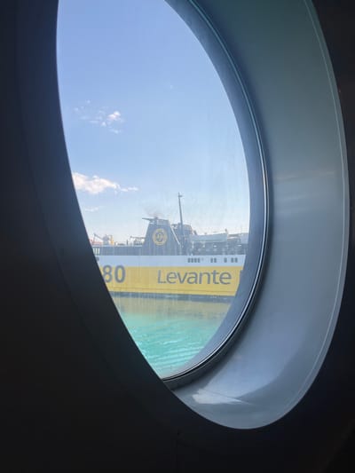 A wide-angle, eye-level shot, taken through a round porthole window on a ferry, captures a view of the port of Kyllini, Greece. The ferry "Levante" is prominently featured, its yellow and white hull marked with the number "80". The ship's black funnel emits a plume of smoke against a clear blue sky dotted with small white clouds. The turquoise water of the sea separates the ferry from the viewer's perspective. The interior of the ferry, including railings and lifeboats, is visible on the upper deck. The porthole's inner frame is a light gray, contrasting with the dark gray of the interior of the room from which the photo is taken. The lighting suggests a bright, sunny day.