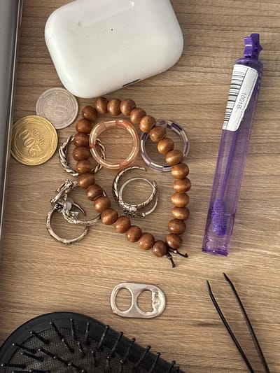 The image displays a collection of personal items scattered on a wooden surface. In the upper portion of the image, a white wireless earbud charging case is visible. To its left, two coins are present: one gold-colored coin with the numeral "200" and another silver-colored coin with the numeral "10". Below these coins, a wooden bead bracelet is arranged in a curved pattern. Several rings are interspersed with the bracelet, including a translucent pink ring, a purple and clear striped ring, and several silver-toned rings with intricate designs. To the right of the bracelet, a purple cosmetic applicator in a clear tube is positioned vertically, featuring a barcode label. In the lower portion of the image, a metal pull tab from a beverage can lies flat. A black hairbrush with numerous bristles is partially visible on the bottom left, and a pair of black tweezers is situated on the bottom right. The overall scene suggests a casual arrangement of everyday objects, and the wooden surface implies an indoor setting, possibly a desk or table. The location is not discernible from the provided image.