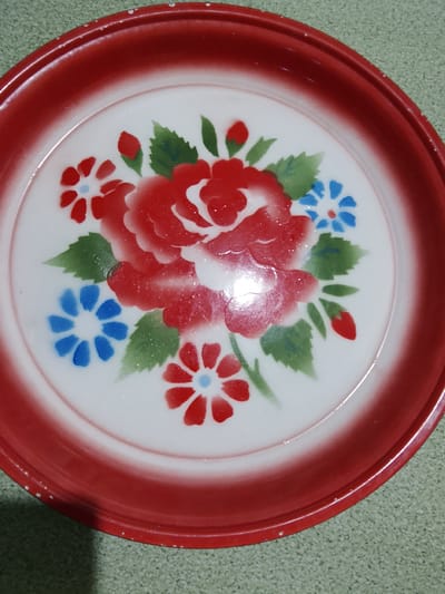 A vintage, red and white enamel plate with a floral design is pictured on a speckled green surface. The plate features a central red rose surrounded by green leaves and smaller blue and red flowers. The red rim of the plate shows some minor wear and chipping. The image is a close-up, top-down view of the plate. The location in Merbopuntong, Indonesia is not depicted or suggested by the image itself.