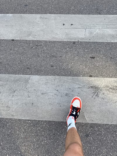 An overhead shot shows a person's right foot, wearing a red and white sneaker and a white sock, stepping onto a crosswalk. The crosswalk consists of alternating wide white stripes and dark gray asphalt. The image is devoid of any specific landmarks or context to place it within Białystok, Poland, or any other city. The lighting suggests it is daytime.