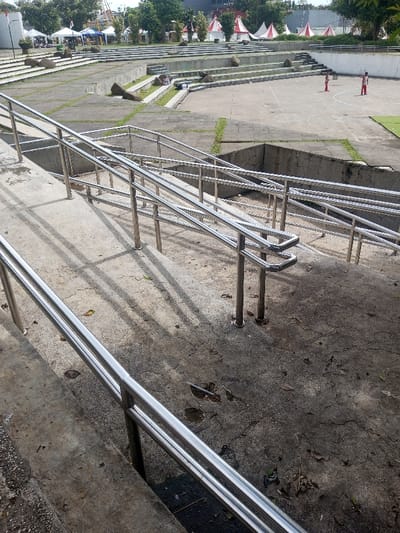 A multi-tiered concrete amphitheater is featured. Atop the tiers, tents and trees are visible. Stainless steel handrails with vertical supports traverse the concrete steps. Two people, likely children, are visible on one of the lower tiers. The handrails are reflective, casting strong shadows on the weathered concrete. The ground shows signs of wear, with scattered debris and discoloration.
