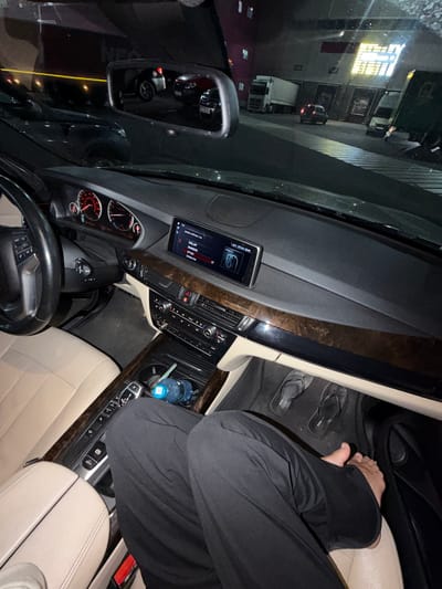 The image shows the interior of a car, with a person's legs and feet visible. The dashboard features a navigation screen displaying "www.silver.ru" and various radio stations. The person is wearing black pants and flip-flops, and their right foot is resting on the accelerator pedal. In the background, visible through the windshield, are trucks and buildings, suggesting a commercial or industrial area, possibly at night. The car appears to be parked or stopped. There are no discernible details to indicate the specific location in Sluchainyi, Russia, beyond the general urban/industrial setting.