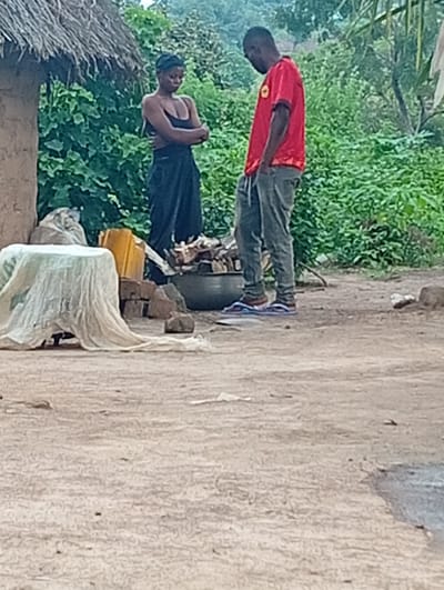 In Tse Igbudu, Nigeria, a Black woman wearing a black spaghetti strap top and a long black skirt stands with her arms crossed. Next to her, a Black man in a red t-shirt and gray pants stands with his hands in his pockets, facing away from the camera. They are in an outdoor setting with a thatched-roof structure to the left and lush green foliage in the background. In the foreground, a table covered with a light-colored cloth, a yellow plastic container, bricks, and a large pot are visible. The ground is made of dirt.