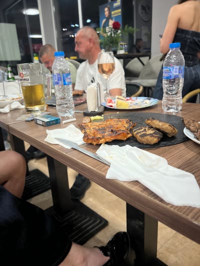 The image depicts a group of people seated at a table in what appears to be an outdoor dining area. The table is laden with food and drinks, including a pitcher of beer, several bottles of water, a plate of grilled meat, and a slice of cake. Two men are visible in the foreground, one of whom is wearing a white t-shirt and the other a green t-shirt. A woman is partially visible in the background, wearing a dark top. The scene is likely set in the city of Zlataritsa, Bulgaria, given the context of the photograph. The overall atmosphere is casual and social, with individuals enjoying a meal together.