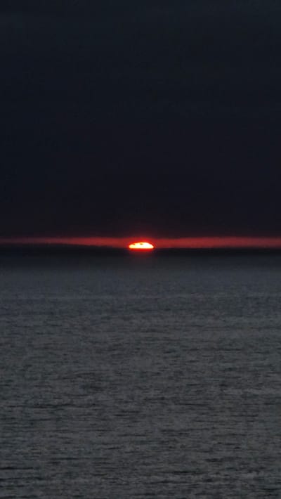 The image shows a sunset over the ocean. The sun is a bright orange disc low on the horizon, casting a red glow across the sky. The water is dark and choppy, with small waves. The sky above the horizon is a deep, dark blue, almost black. There are no people or man-made objects visible in the image. This appears to be a natural scene, possibly taken from a beach or a cliff overlooking the ocean. Given the location of Encinitas, a coastal city in San Diego County, California, this sunset scene is likely from the Pacific Ocean, west of the city.