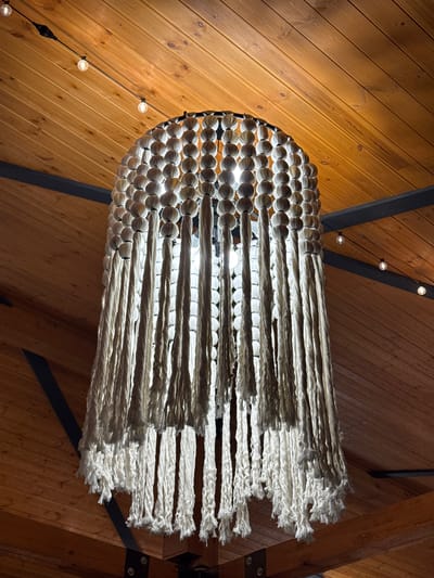 A close-up, low-angle interior shot shows a macrame chandelier with a white, spherical beaded canopy and long, cream-colored fringe. The chandelier is suspended from a wooden plank ceiling and is illuminated by warm, ambient light. The ceiling is made of narrow, parallel wood planks, and several small, bare incandescent bulbs on a black wire string are visible against the wood. Dark metal beams are also visible, supporting the ceiling structure. The setting appears to be a restaurant or a similar establishment in Cholpon-Ata, Kyrgyzstan, given the context provided by the prompt. No people are visible in the image.