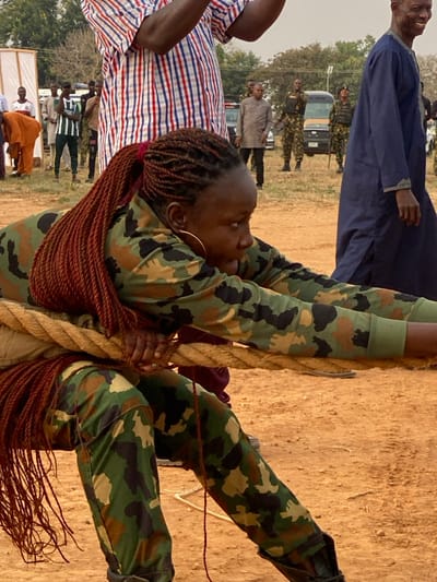 A woman with red braided hair is climbing a thick rope, wearing camouflage clothing and gold hoop earrings. She is surrounded by onlookers in a dry, dusty outdoor setting. In the background, there are people wearing various clothing, including a blue robe and casual shirts, a parked car, and uniformed individuals standing nearby. The image is slightly rotated counter-clockwise.