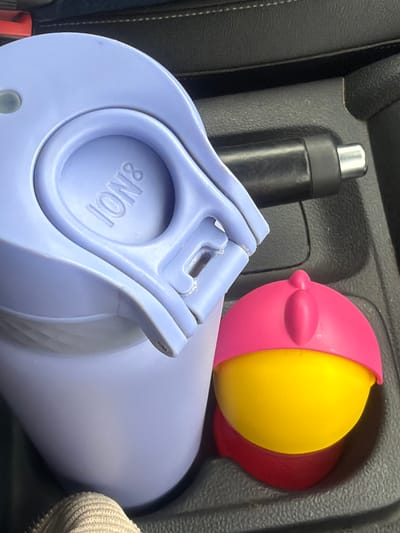 The image shows the interior of a car. In the center console, there is a black travel mug with a silver lid, a large, light purple water bottle with an "ION8" brand marking on the lid, and a toy ball. The toy ball is segmented into pink, yellow, and red sections. The car interior appears to be dark. The image was not taken in Kamburovo, Bulgaria.