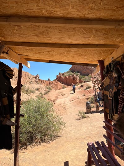 A view from inside a structure, possibly a yurt or open-sided building, looks out onto a landscape in Tosor, Kyrgyzstan. The interior walls of the structure are constructed of light-colored wood. Hanging inside are decorated saddles and other equestrian equipment. Outside, the landscape is arid and consists of red rock formations and sparse vegetation, including a large bush in the foreground. In the middle distance, a person walks along a path. The sky is blue and clear.