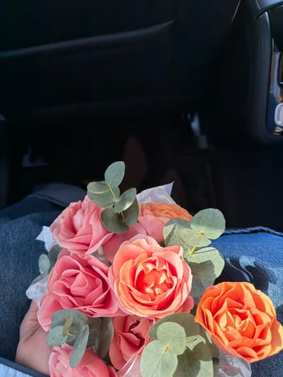 A person is seated inside a vehicle, holding a bouquet of flowers on their lap. The bouquet consists primarily of light pink and orange roses, interspersed with green eucalyptus leaves and white tissue paper. The hand holding the bouquet, visible on the lower left, has light skin tone. The person is wearing blue denim jeans, partially visible on their lap. The background shows the dark interior of a car, specifically the dashboard or passenger footwell area, with various dark gray and black surfaces and faint reflections of interior components. Sunlight illuminates the flowers and denim, casting distinct shadows. The specific location within the city of Kukui, Russia, cannot be determined from the visual information provided in the image.
