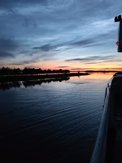 The image depicts a sunset over a body of water, possibly a river or lake, with a shoreline lined with trees. The sky is filled with dramatic clouds painted in hues of blue, grey, and orange, indicating the last moments of daylight. The water reflects the sky and the treeline, creating a mirror image. In the foreground, the edge of a boat or vessel is visible on the right side of the frame. The water exhibits ripples, suggesting movement. On the distant shore, a few structures, including what appears to be a crane or industrial equipment, are silhouetted against the sunset. The overall atmosphere is serene and tranquil. The location is identified as the city of Sheksna, Russia.