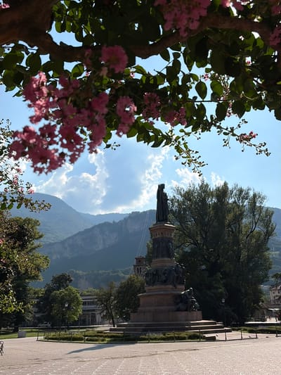 This is a vertical, eye-level, medium shot of a park area with a monument and a mountain range in the background. The scene is framed by the branches of a tree with pink flowers and green leaves in the foreground on the left. The monument, a large bronze statue on a stone pedestal, is located in the middle ground to the right. A paved walkway is visible in the far right of the image, with a manicured garden area to its left. The background features a lush green mountain range under a bright blue sky with scattered white clouds. The lighting suggests it is daytime, likely mid-morning or afternoon, with the sun casting shadows. No people are clearly visible, but the presence of the monument and park implies a public space. The overall mood is serene and picturesque, highlighting the natural beauty and the monument. No text is visible in the image. The location context is Trento, Italy.