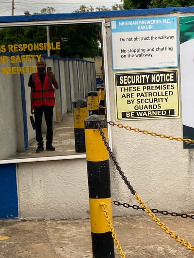 A man wearing a red safety vest and black pants stands in front of a reflective surface, taking a photo with his phone. He is holding a black object in his left hand. Behind him, in the reflection, is a pathway lined with concrete pillars. To the right of the man, there are two signs. The top sign, in blue and white, reads "NIGERIAN BREWERIES PLC, KAKURI" and "Do not obstruct the walkway. No stopping and chatting on the walkway." Below that, a yellow and black sign states "SECURITY NOTICE THESE PREMISES ARE PATROLLED BY SECURITY GUARDS BE WARNED!". Chains are draped across the foreground, attached to bollards that are painted with yellow and black stripes. The environment appears to be outdoors, with trees visible in the reflection.