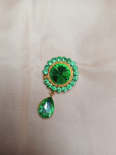 The image displays a single piece of ornate costume jewelry, resembling a brooch or pendant, positioned against a light beige fabric background with subtle texture and folds. The item is photographed in an indoor setting; the specified location of Qarabucaq qəsəbə, Azerbaijan is not visually depicted within the image.
The jewelry consists of a primary circular element and a dangling teardrop-shaped component. The circular element features a prominent, faceted, dark green, round gemstone at its center, surrounded by a textured gold-colored bezel. This central stone is encircled by approximately 20 smaller, faceted, lighter green, round gemstones set in a gold-colored frame, forming a floral or sunburst pattern. Attached to the bottom of the circular element via a small gold-colored jump ring is a single, faceted, lighter green, teardrop-shaped gemstone, also set in a gold-colored bezel. No individuals, actions, or direct interactions are present in the scene.