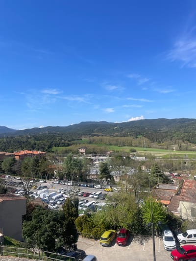 The image shows a landscape taken from an elevated position in Hostalric, Spain. The left side of the image primarily consists of a clear blue sky with wisps of white clouds.
In the center of the image, a mountain range is visible, covered in lush green vegetation. At the foot of the mountains, there is a flat green field, potentially a meadow or farmland.
On the right side of the image, a densely populated area is seen. There are numerous buildings with red-tiled roofs. A parking lot filled with various cars, predominantly white, is present. Trees and greenery are interspersed among the buildings and parking area.