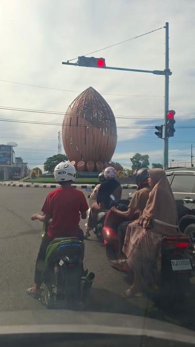 In the city of Sigli, Indonesia, a street scene unfolds at a traffic intersection. The dominant landmark is a large, reddish-brown, dome-shaped sculpture with an intricate lattice pattern, resembling a giant nut or seed pod. Traffic lights are visible, with the top light in one signal displaying red. Several motorcycles are stopped at the intersection. On the nearest motorcycle, a person wearing a red shirt and a white helmet is viewed from behind. Behind them, two other motorcycles carry passengers, all wearing helmets and headscarves. The scene is set against a bright sky with scattered clouds and features utility poles and wires. Signs and buildings are present in the background, suggesting a commercial area.