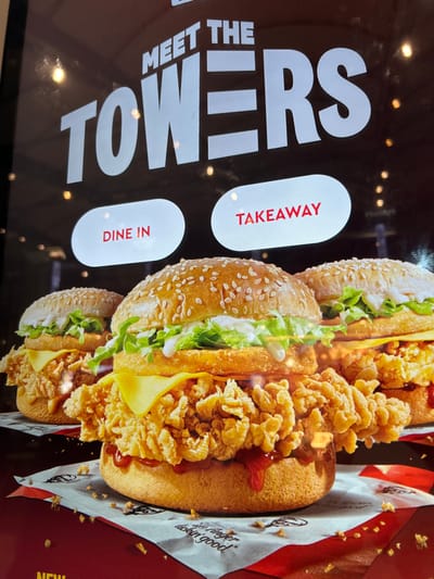 This image is an advertisement, likely displayed on a digital screen within a fast-food restaurant, possibly KFC, given the branding. The overall presentation is rotated 90 degrees clockwise.

The main subjects are several long, bun-based food items referred to as "The Towers." These appear to be variations of a fried chicken sandwich or hot dog-style bun, featuring crispy, irregular pieces of fried chicken, melted yellow cheese, fresh green lettuce, and a red sauce (possibly ketchup or a spicy condiment). The buns are light brown and covered with sesame seeds. The food items are arranged dynamically, appearing to be stacked or layered within the advertisement.

The background is predominantly dark with blurred, circular lights, creating a bokeh effect, suggesting an indoor setting. To the right, a reddish-brown area includes crumpled paper with the iconic KFC logo and the slogan "finger lickin' good" printed on it, reinforcing the brand.

Visible text on the left, rotated, reads "MEET THE TOWERS" in large white capital letters. Below this are two white, oval-shaped interactive elements or buttons, also rotated, with red text: "TAKEAWAY" and "DINE IN." A partially visible "NEW" in white text against a red background can be seen at the bottom right.

The image depicts a promotional display for new menu items, allowing customers to choose between ordering for dine-in or takeaway, within a fast-food context in Agios Dimitrios, Greece.