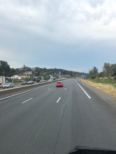 The image shows a view from a vehicle traveling on a highway in Ternay, France. The highway has multiple lanes, indicated by white lane markings. On the left side of the highway, there is a concrete barrier, and beyond that, a train track and some buildings are visible. On the right side of the highway, there is a grassy verge with trees and dry vegetation. Several vehicles, including cars and trucks, are present on the highway, traveling in the same direction as the viewpoint. In the distance, there is a town with buildings and a church or castle on a hilltop. The sky is overcast with clouds. A red car is prominently featured in the foreground on the highway. Road signs with French text are also visible.