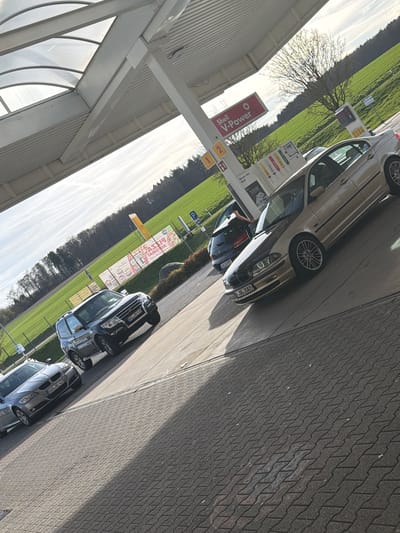 The image captures a Shell gas station in Lautzenhausen, Germany, with several parked cars. The gas station features a Shell V-Power sign and gas pumps. Three cars are visible from the end. A silver car is parked at the first pump and someone is fueling their car. Next is a black car. Further down is a light blue car. Last is a silver car parked on the end. The environment consists of a green field surrounding the gas station and a paved area.