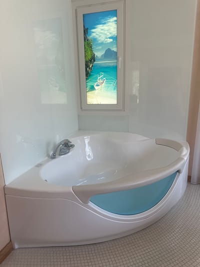 This image depicts a modern bathroom with a corner bathtub. The bathtub is white with a light blue accent panel and is positioned in a corner of the room. Above the bathtub, a small window is visible, featuring a scenic image of a tropical beach with boats. The walls are white and appear to be made of a smooth, reflective material. The floor is tiled with small, square beige tiles. To the right of the bathtub, a light-colored curtain is partially visible. There are no people or animals in the image. The scene is well-lit and suggests a clean, spa-like environment. The location of this bathroom is not specified in the image or its context.