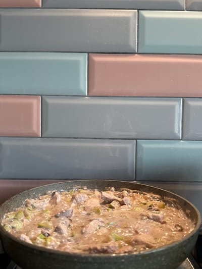 A close-up, eye-level shot shows a cooking pan on the right side, filled with a creamy stew. The stew contains visible pieces of meat and green vegetables, and it is bubbling, indicating it is actively cooking. The pan has a speckled gray exterior. The background on the left consists of a tiled wall. The tiles are rectangular and arranged in a subway pattern, with alternating colors of light gray, pale pink, and light blue. The grout between the tiles is dark gray. The lighting suggests an indoor setting, likely a kitchen, with no indication of the time of day or weather. There is no visible text in the image. The scene captures a moment of food preparation, with the focus on the bubbling stew and the textured tiled background.