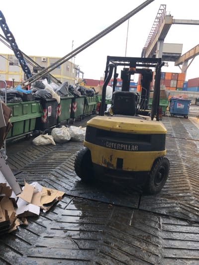 The scene depicts a section of the Port of La Spezia, Italy. A yellow and black Caterpillar forklift, facing away from the viewer, is positioned in the midground on a wet, dark, grooved metal deck. To the left, a row of interconnected green industrial waste bins is overflowing with black and white refuse bags. Several white sacks are lying on the wet deck in front of these bins. Pieces of flattened cardboard boxes are scattered on the deck in the lower-left corner. Heavy metal chains and cables descend diagonally from the upper left. In the background, modular buildings are visible behind the waste bins. To the right, a large gantry crane overlooks a dense arrangement of stacked, multi-colored shipping containers. A blue wheeled dumpster is also visible in the right background. The sky is overcast. No individuals are present.