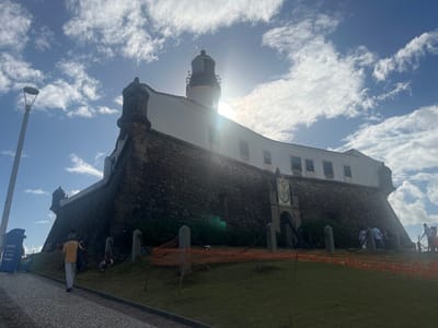 The image displays the Forte de Santo Antônio da Barra and the Farol da Barra (Barra Lighthouse) in Salvador, Bahia, Brazil, under a bright blue sky with scattered white clouds. The sun is positioned behind the lighthouse, causing significant backlighting and lens flare.
The fort consists of a robust, dark stone base with discernible corner bastions or turrets. A lighter-colored heraldic crest is affixed to the stone wall's central section. Above the stone base, the fort transitions into a white building featuring multiple rectangular windows. Capping the entire structure is the Farol da Barra, a black and white striped lighthouse tower with a black domed top.
In the foreground, a patterned cobblestone pathway curves upwards from the lower-left. A lone individual, viewed from behind, wearing a yellow shirt and light-colored pants, walks along this path. To the immediate left of the path stands a tall, grey streetlamp alongside two blue waste bins. The ground leading up to the fort is a green grassy slope, partially delineated by a horizontal orange safety net. Several individuals are present on this slope, some seated or resting near the fort's base. Another group of people is visible near a partially obscured entrance in the fort's stone wall on the right side of the image.